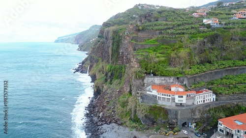 Dramatic coastal cliffs of Madeira, Portugal, with lush vegetation and a waterfall cascading into the Atlantic. Overcast skies enhance the moody, rugged beauty.