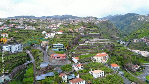 Aerial view of a Ponta do Sol village cascading down lush, terraced hillsides in Madeira, Portugal. Traditional homes blend with modern infrastructure under an overcast sky.
