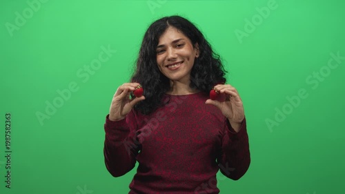 Young arab brunette woman holding strawberries between fingers and puckering lips in green studio; playful flirtation.