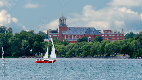 Red sailboat on a waterway on a summer  day with a tree-lined shore in the background.