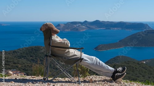 Hiker relaxing on camping chair and enjoying scenic sea view. Scenic view of a man sitting in a camping chair on a cliff, enjoying the stunning seascape with islands and mountains on a sunny day