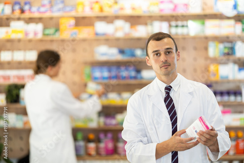 Kind young apothecary holding a box with remedy standing against shelves full of medicine in drugstore