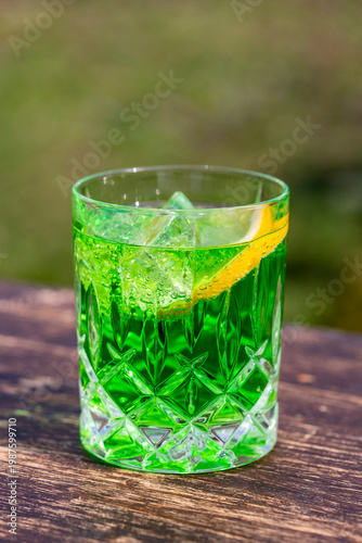 Refreshing green cocktail with ice and lemon in crystal glass. Cold green soda or cocktail with ice cubes and lemon slice in glass on wooden table. Bright summer drink with blurred outdoor background