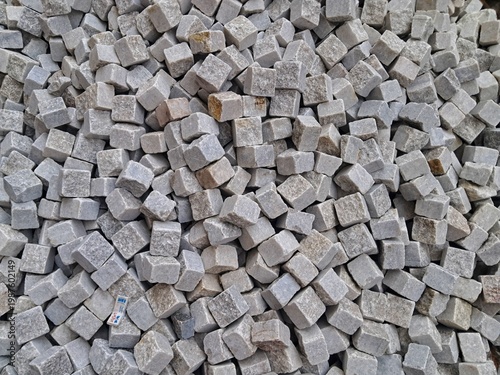 A top-down close-up view of a pile of gray granite paving stones, showing the natural texture and square shapes used for traditional European streets.