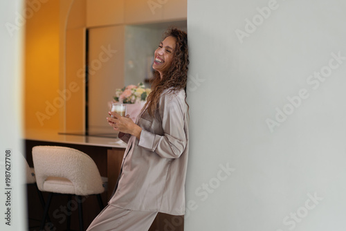 Cheerful woman with curly hair in silk pajamas leaning on wall and holding glass of coffee. Beautiful lady is enjoying morning refreshment in modern house