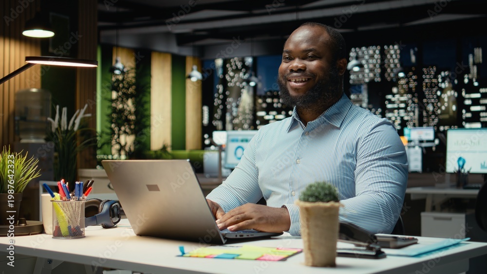 Fototapeta premium Portrait of african american joyful guy manages emails and administrative tasks, documenting insights for a multinational company. Meeting business objectives late at night in office.
