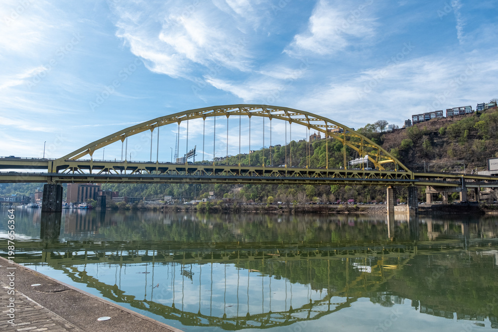 Obraz premium Fort Pitt Bridge Crosses Monongahela River in Pittsburgh With Clear Sky and Green Hillside in View During Daylight Hours