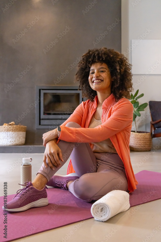 Naklejka premium African American woman in coral jacket sitting on purple mat at home, holding water bottle, towel