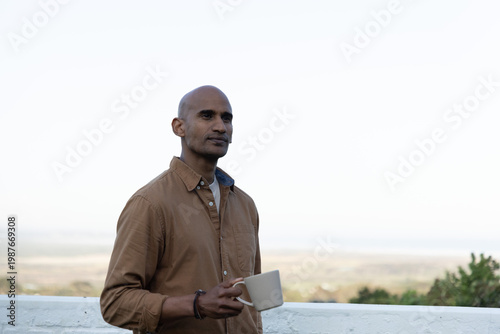 Middle-aged Indian man standing on balcony holding beige mug and wearing brown button-up shirt