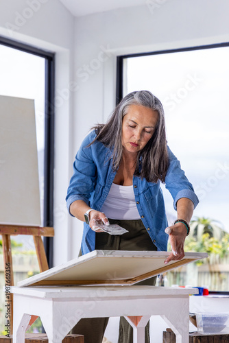 Senior woman smoothing canvas using palette knife at white table near windows showing palm tree
