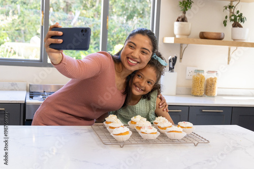 Indian mother and child posing smartphone selfie in kitchen by frosted cupcakes, wearing blue bow