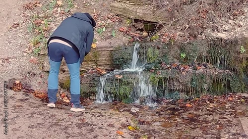 Individual in casual attire bends down to collect water from a small stream surrounded by autumn foliage and rocky terrain in a natural outdoor setting
