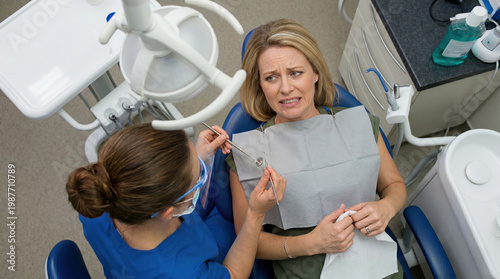An apprehensive woman winces in a dental chair while a dentist holds a mirror and probe. High-angle view of a clinical setting showing dental equipment, medical tools, and a nervous female patient.