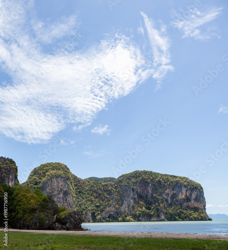 Tropical beach with cliffs at Hat Ratcha Mongkhon, Sikao, Trang, Thailand. Clear blue sky over green hills and shoreline. Calm waters and lush vegetation on rocky landscape.