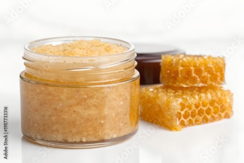 Natural honey-infused exfoliating scrub in a glass jar, accompanied by raw honeycomb pieces on a pristine white background, emphasizing organic skincare and a wholesome beauty routine