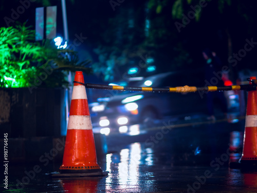 Traffic cone and barrier on wet city street at night with blurred headlights, reflecting rain-soaked pavement and urban safety control in a moody nighttime scene.