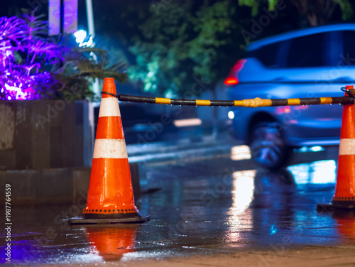 Traffic cone and barrier on wet city street at night with blurred headlights, reflecting rain-soaked pavement and urban safety control in a moody nighttime scene.