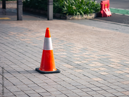 Single orange traffic cone placed on paved sidewalk, marking caution area in an urban setting, highlighting safety, control, and temporary restriction in public space.