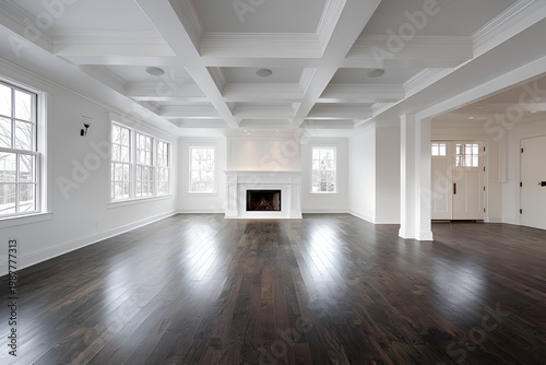 A spacious, bright interior with gleaming hardwood floors, white walls, and a coffered ceiling. A fireplace is centered. Large windows allow natural light