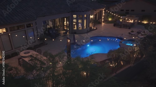 A 4k night aerial view of a desert landscaped home in Arizona featuring a travertine tiled pool deck with an outdoor fireplace and kitchen.