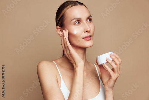 Tableau sur toile Woman applying skincare cream to face, holding small jar, using cotton pad, calm