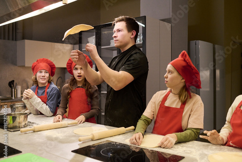 Quadro su tela Young adult Caucasian man teaching children tossing pizza dough in cooking class, learning teamwork and food skills