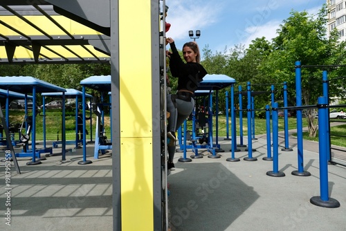 A young woman on an outdoor climbing wall in a city park. The training takes place on a modern workout area with professional equipment.
