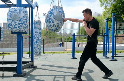 An energetic middle-aged man is practicing a straight punch on a punching bag. The training takes place outdoors on a modern workout area. The atmosphere is one of active healthy living