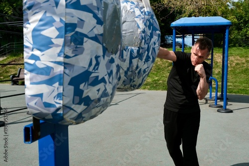 An energetic middle-aged man is practicing a straight punch on a punching bag. The training takes place outdoors on a modern workout area. The atmosphere is one of active healthy living