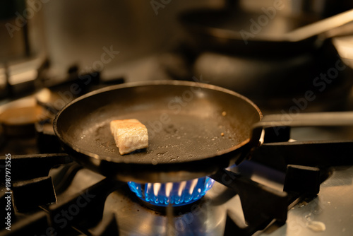 Frying pan gas stove cooking fish kitchen flame skillet burner seafood saute pan gas stove burner with blue flame, small fish piece sizzling oil, warm indoor light, home kitchen meal preparation