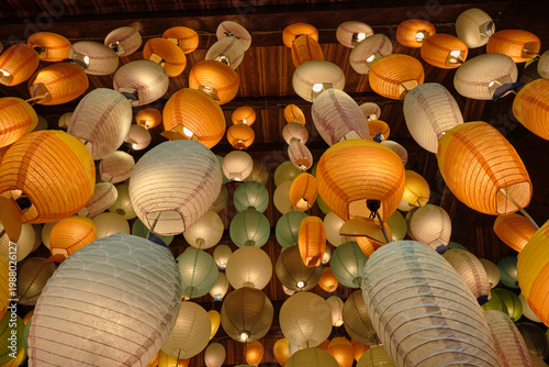 A closer, low-angle perspective looking up at glowing paper lanterns in shades of blue, green, orange, and cream with visible wooden structure.