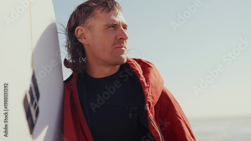 Surfer leans against board, gaze fixed on horizon. Red jacket contrasts with bright sky and calm sea. He appears thoughtful, waiting for the next wave