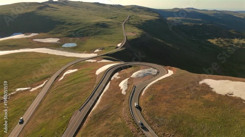 Beartooth Highway in Montana winds through alpine terrain with dramatic hairpin curves. Cars travel along the scenic mountain pass with patches of snow and green meadows visible from above.