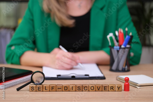 A person sits at a wooden desk, writing intently on a clipboard. Nearby, wooden blocks spell out self-discovery, reflecting a journey of personal growth amidst colorful pens and tools.