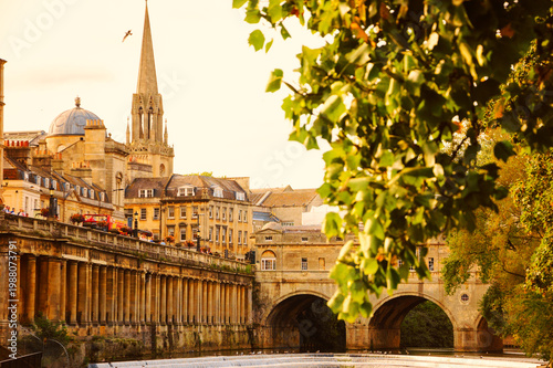 Pulteney Bridge over the River Avon in Bath, UK, historic cityscape with St Michael's Church glowing in golden sunset light, framed by blurry tree leaves and classic Georgian architecture