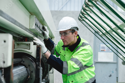 Caucasian man operates a corrugated cardboard conveyor machine in a dimly lit paper factory, wearing a white helmet and dark work clothing for industrial inspection and quality control.