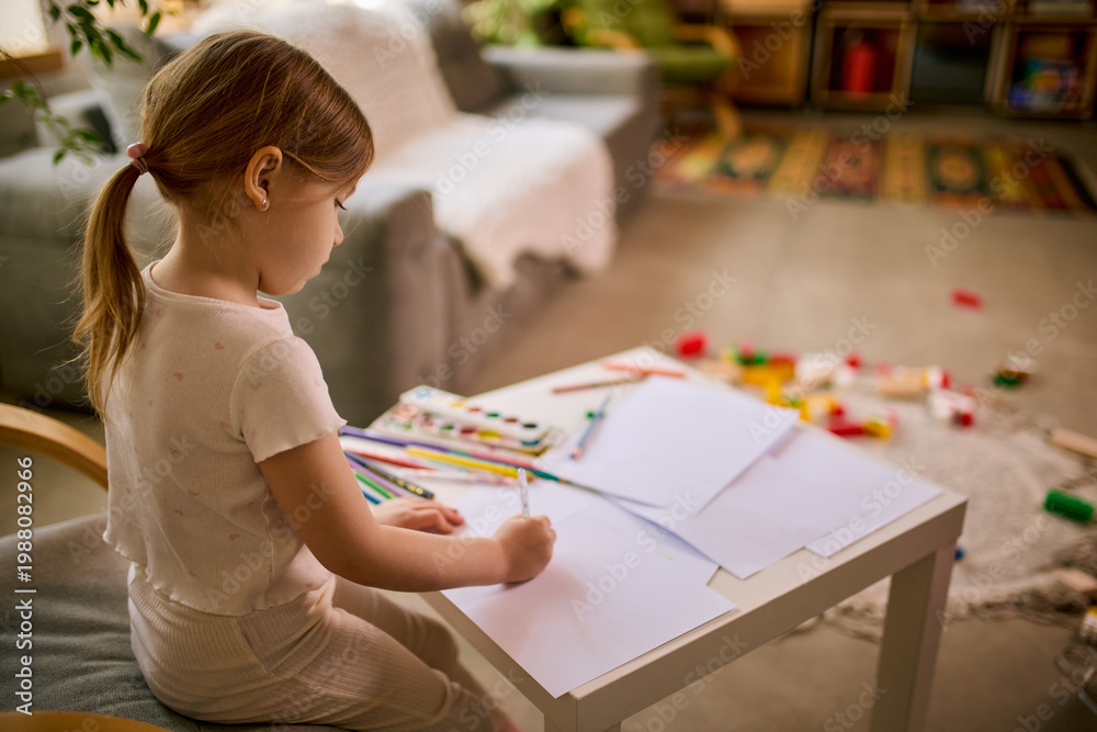 Fototapeta premium Little girl drawing with pencils at small table in cozy home interior. Concept of kids products promotion, creative classes advertising, early education materials and hobby development content.