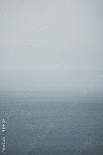 Minimalist view of a calm sea surface disappearing into thick white fog. Misty ocean horizon under a grey overcast sky.