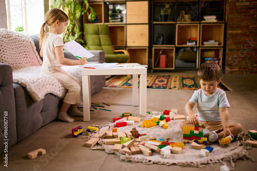 Little children playing separately at home with girl drawing and boy building blocks on floor. Concept of parallel play behavior, sibling independence and home activity routines.