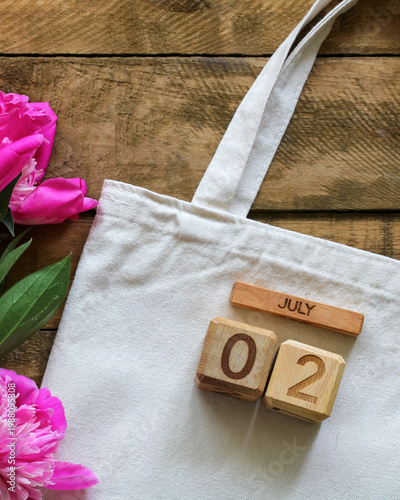 July 2 calendar date with eco tote bag and pink peony flowers on wooden background