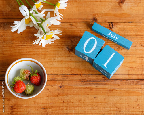 Top view of a July 1 calendar made of wooden blocks with fresh strawberries in a bowl and chamomile flowers on a rustic wooden table