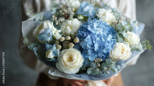 a bouquet of blue hydrangeas and white roses, wrapped in paper and held by an asian woman wearing casual attire. the background is gray with soft lighting, creating a romantic atmosphere. it was taken
