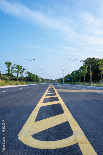 New Asphalt Road with Bold Yellow Centerline, Streetlights, and Lush Greenery Under Blue Sky