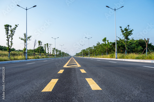 Empty Modern Asphalt Highway with Yellow Centerline, Streetlights, and Green Verges Under Clear Blue Sky