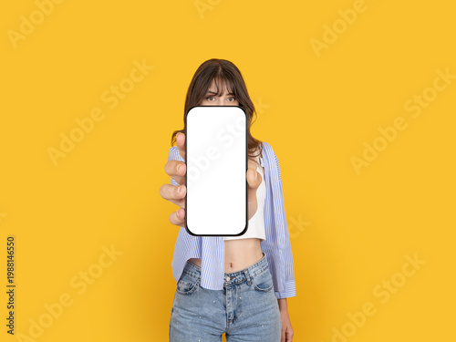 Obraz na plátně Caucasian woman holding smartphone toward camera with blank screen mock up, covering face partially, presenting mobile app, wearing striped shirt, isolated on yellow background with copy space