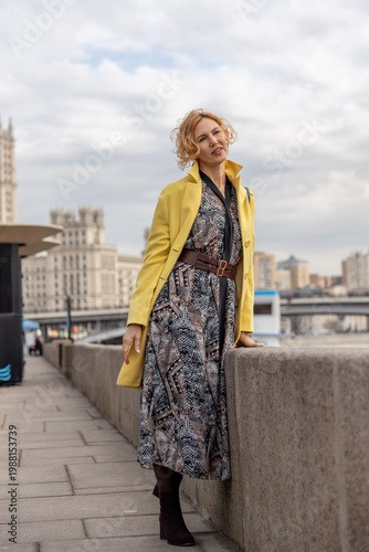 Elegant woman in patterned dress and yellow coat posing at city embankment