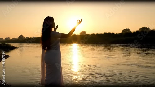Woman in white dress praying by the river at sunset.
