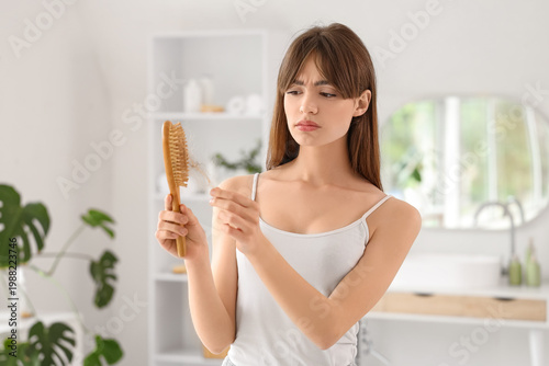 Beautiful young shocked woman holding brush with fallen down hair in bathroom...