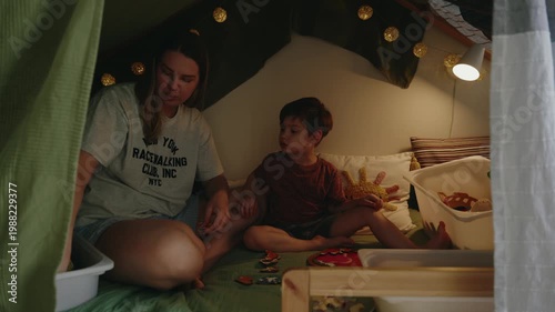 Mother and son engage in playful activities on a bed inside a cozy fort, surrounded by toys and soft lighting creating a warm atmosphere