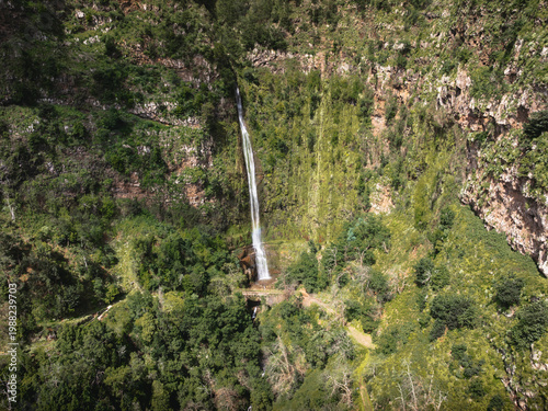 Aerial view of the Cascata do Monte waterfall cascading down a lush green cliffside towards the historic Ponte do Pisao stone bridge in Monte, Madeira, Portugal.